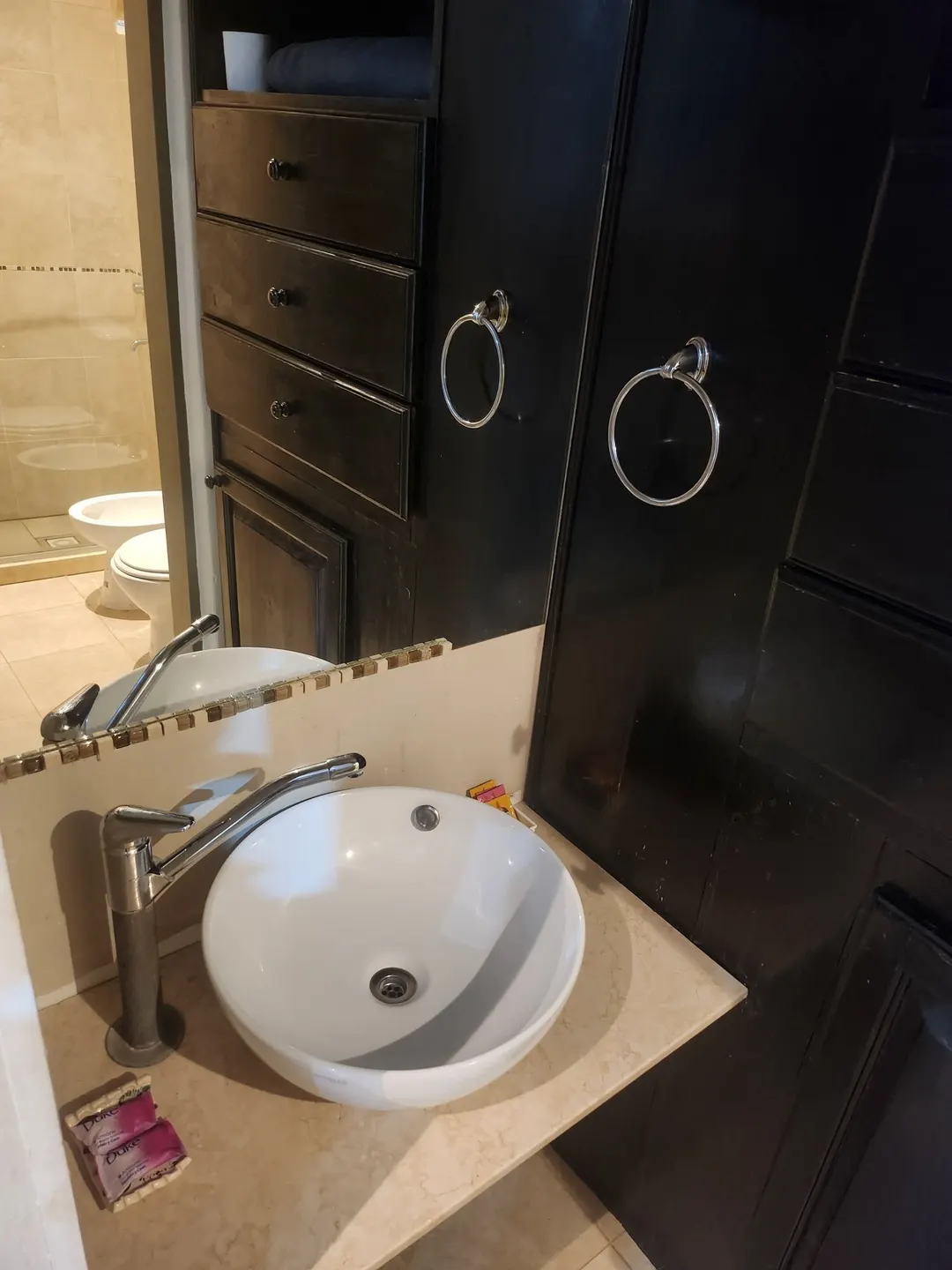 Lavabo blanco sobre mesada clara, espejo y mueble oscuro, con la ducha al fondo