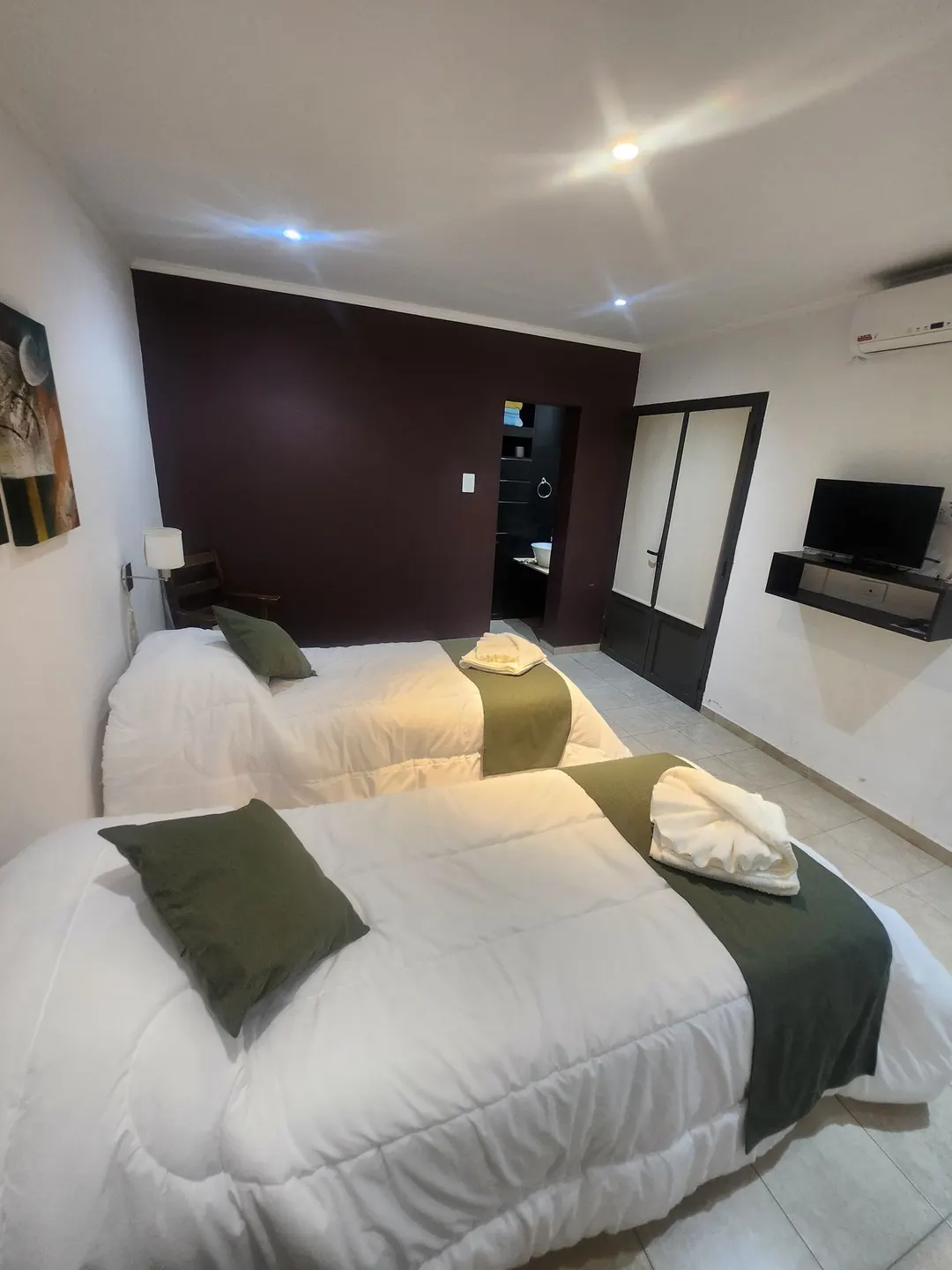 Dos camas individuales con detalles verdes, televisor y puerta del baño al fondo