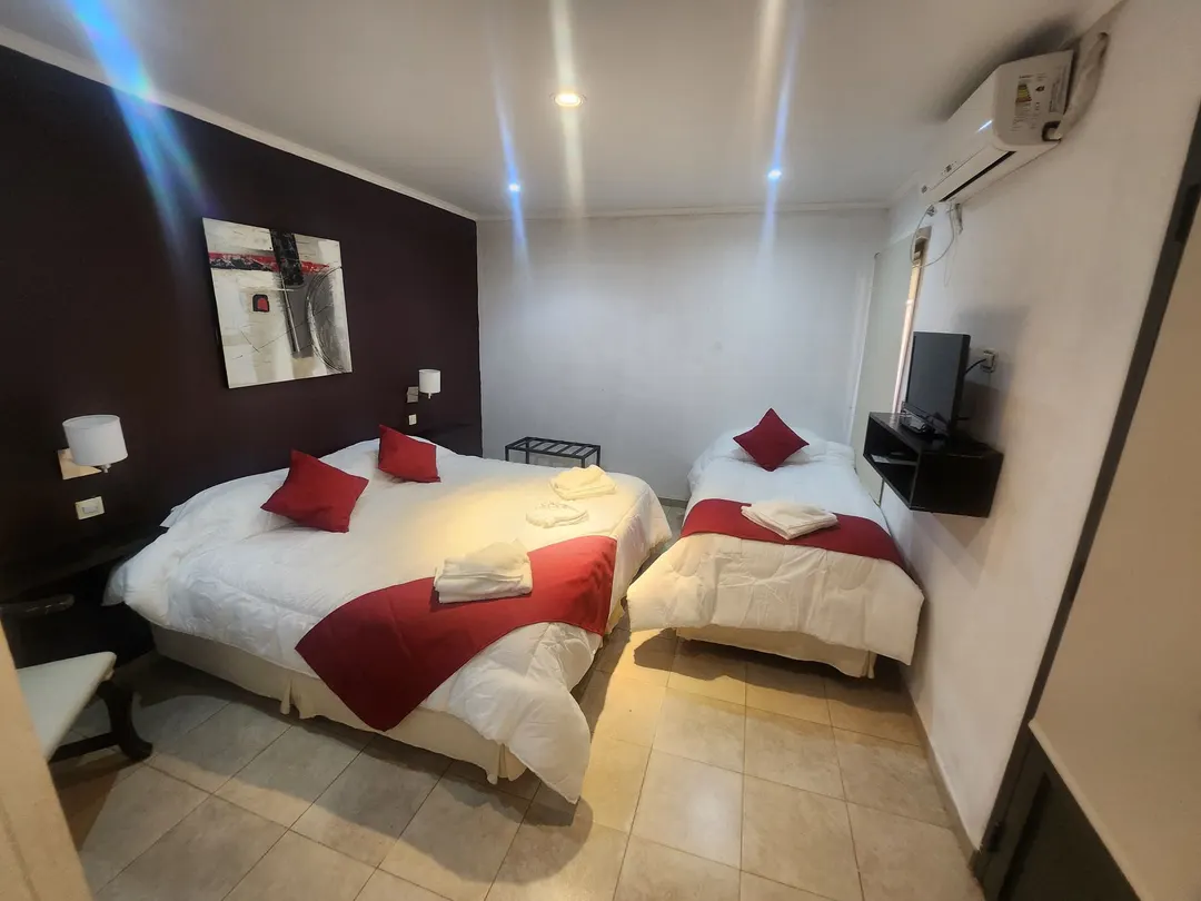 Tres camas individuales, dos juntas, con almohadones rojos, TV y aire acondicionado